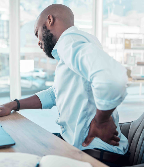 A man at a desk holding his lower back in discomfort.